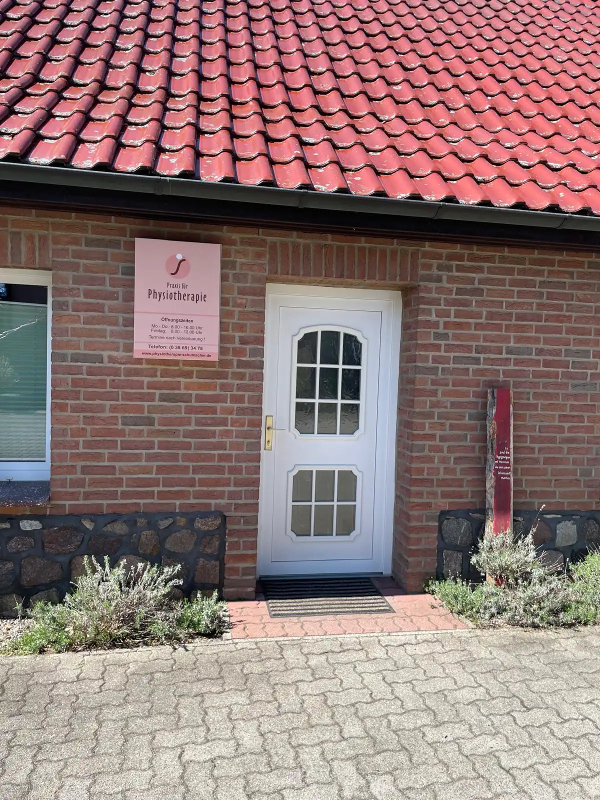 Eingangstür mit weißem Rahmen auf rotem Backsteingebäude, darüber ein Schild mit der Aufschrift „Physiotherapie Schumacher & Hagen“. Dach mit roten Ziegeln, davor ein Gehweg mit Pflanzen.