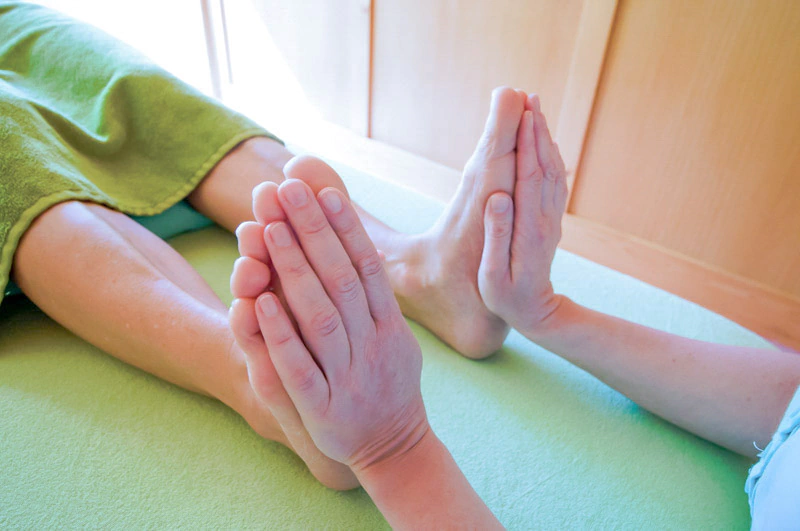 Handhaltung als Teil einer physiotherapeutischen Anwendung bei der Physiotherapie Schumacher & Hagen. Eine Person hält die Füße eines liegenden Patienten auf einer grünen Liege, möglicherweise bei einer Mobilisationstechnik.