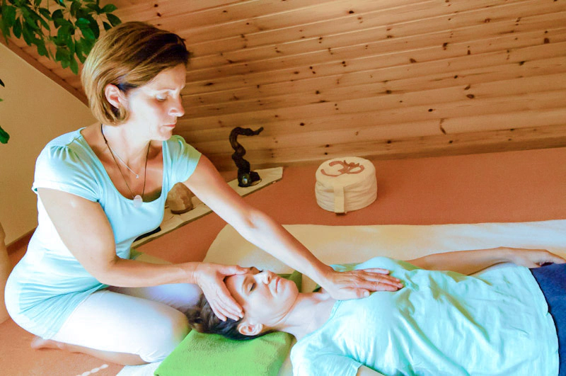 Eine Therapeutin führt eine manuelle Therapie durch, indem sie sanft Kopf und Brust einer liegenden Person berührt. Im Hintergrund ist eine Holzdecke zu sehen. Kontext: Physiotherapie Schumacher & Hagen.