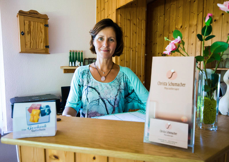 Eine Person steht hinter einem Empfangstresen in der Physiotherapie Schumacher & Hagen. Auf dem Tresen befinden sich ein Schild mit der Aufschrift „Christa Schumacher Physiotherapie“ und eine Blumenvase. An der Wand ist ein Apothekerschrank erkennbar.