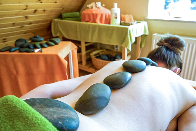Eine Person liegt mit erhitzten Steinen auf dem Rücken, die eine Hot-Stone-Massage im Rahmen der Physiotherapie Schumacher & Hagen zeigen. Im Hintergrund ist ein Behandlungsraum mit Tischen, die mit Tüchern abgedeckt sind, sowie ein Regal mit Massagezubehör.